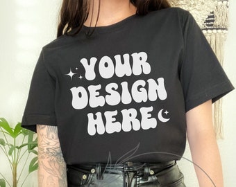 May include: A black t-shirt with the text "YOUR DESIGN HERE" in white, retro-style lettering. The shirt is being worn by a person with tattoos on their arm.