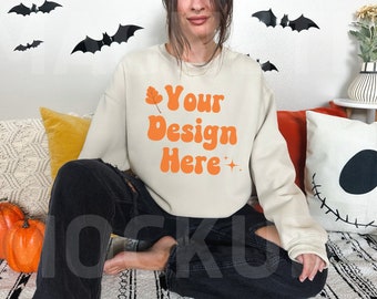 Puede incluir: Una mujer lleva una sudadera de color crema con el texto "Your Design Here" en naranja. Está sentada en una alfombra estampada con calabazas y una almohada con la cara de Jack Skellington en blanco y negro.