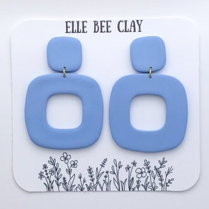 Peut inclure: Paire de boucles d'oreilles carrées bleu clair, avec un petit carré en haut, reliées par des crochets dorés. Elles sont présentées sur une carte blanche avec le texte "ELLE BEE CLAY" et une illustration florale.