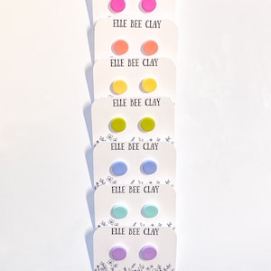Peut inclure: Six paires de boucles d'oreilles clous en argile colorées présentées sur des cartes blanches avec le texte "ELLE BEE CLAY". Les boucles d'oreilles sont dans des tons de rose, orange, jaune, vert, bleu, turquoise et violet.