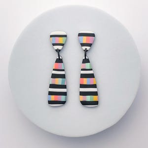 May include: A pair of handmade earrings with a striking design. Each earring has a black and white striped pattern with rainbow-colored sections. The earrings are teardrop-shaped and presented on a pale blue surface.