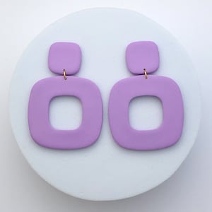 Peut inclure: Une paire de boucles d'oreilles géométriques de couleur lavande. Chaque boucle d'oreille présente un petit carré supérieur relié à une forme carrée ouverte plus grande en dessous. Les boucles d'oreilles sont présentées sur une surface circulaire blanche.