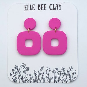 May include: A pair of bright pink, square-shaped earrings with a circular top, connected by a gold-colored hook. The earrings are displayed on a white card with the text "ELLE BEE CLAY" and a floral design.