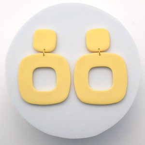 May include: A pair of pale yellow square earrings with a smaller square top and a larger square bottom. The earrings are connected by a small gold-colored metal loop. The earrings are displayed on a light blue circular surface.