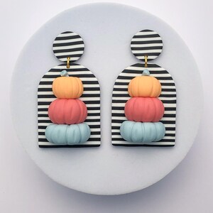 May include: A pair of colorful pumpkin earrings. Each earring features a stack of three pumpkins in orange, pink, and blue, set against a black and white striped background. The earrings are attached to a black and white striped circle.