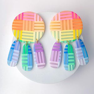 May include: A pair of colorful, abstract earrings. Each earring features a circular top with a woven pattern in shades of pink, orange, yellow, and green. Below, elongated teardrop shapes dangle, with similar color patterns.