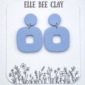 May include: A pair of light blue clay earrings with a square shape and a small circle on top. The earrings are on a white card with the text "ELLE BEE CLAY".