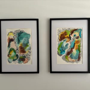 Könnte beinhalten: Zwei gerahmte abstrakte Aquarellbilder mit schwarzen Rahmen. Die Bilder zeigen organische Formen und Linien in Grün-, Blau-, Braun- und Weißtönen.