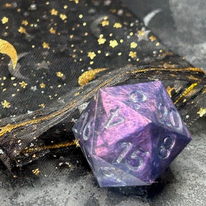May include: A purple and blue iridescent 20-sided die with black numbers. The die is sitting on a black fabric bag with gold glitter stars and moons.
