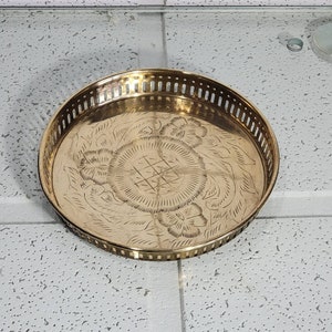Peut inclure: Un plateau rond en laiton avec un motif gravé décoratif de fleurs et de feuilles. Le plateau a un bord festonné et un rebord surélevé.