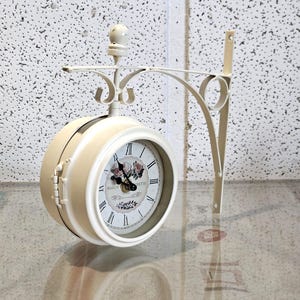 Könnte beinhalten: Cremefarbene, doppelseitige Uhr mit römischen Ziffern und floralen Akzenten. Die Uhr ist an einer dekorativen Metallhalterung aufgehängt. Das Zifferblatt zeigt "MAISON RETTE". Die Uhr hat einen Durchmesser von ca. 20 cm.