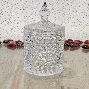 May include: A clear glass jar with a diamond pattern and a decorative lid. The cylindrical jar has a lid with a pointed finial. The jar sits on a reflective surface. Decorative glass beads are in the background.