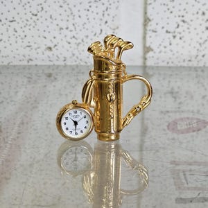Puede incluir: Una bolsa de golf dorada en miniatura con palos de golf y un pequeño reloj redondo. La esfera del reloj es blanca con números y manecillas negras. La bolsa de golf tiene un asa y es muy detallada.