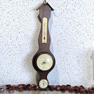 May include: A wooden weather station with a thermometer, barometer, and hygrometer. The thermometer displays both Fahrenheit and Celsius. The barometer has a white face with black lettering. The hygrometer is at the bottom. The wood is a dark brown color.