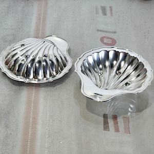 Peut inclure: Deux plats argentés en forme de coquillage. L'un est fermé, l'autre ouvert, révélant l'intérieur. Les deux ont un bord festonné et trois petits ornements ronds. Les plats sont sur une surface réfléchissante.