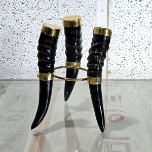 May include: A decorative horn stand featuring three black horns with brass accents. Each horn has a brass cap and is connected by a gold-colored ring. The stand is designed to hold items and adds a rustic touch.
