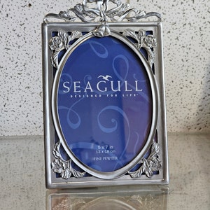 Puede incluir: Un marco de fotos plateado con una abertura ovalada y un diseño floral decorativo. El marco está hecho de peltre fino y tiene las palabras "SEAGULL DESIGNED FOR LIFE" en la parte delantera. El marco mide 5 x 7 pulgadas (13 x 18 cm).
