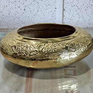 May include: A gold-coloured, oval bowl featuring embossed floral and leaf patterns around the rim. The bowl has a wide opening and a polished surface. The design is symmetrical, with detailed carvings. The bowl is likely made of brass or a similar metal.