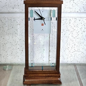 May include: A wooden Bulova clock with a rectangular face and a glass front. The clock features a light-colored square clock face with black hands and the Bulova logo. The frame is a dark brown color with a decorative design.