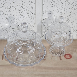 May include: Two clear glass cake domes with intricate designs. The larger dome has a scalloped edge and a heart pattern. The smaller dome has a pedestal base.