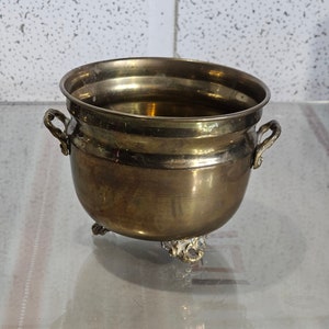 Może przedstawiać: Mały, mosiężny pojemnik z dwoma uchwytami i trzema nóżkami. Pojemnik jest pusty i ma błyszczące wykończenie.