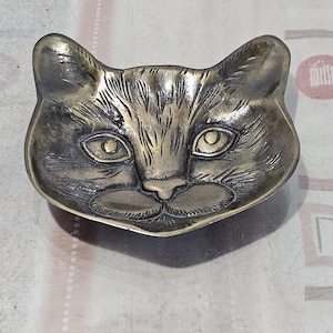 Puede incluir: Un plato de metal para joyas con forma de cara de gato. El plato es de color dorado claro y tiene un diseño detallado de una cara de gato con pelaje, ojos y nariz.