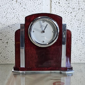 May include: A dark red wooden clock with silver accents and a white face. The clock face has the word "Quartz" and the brand name "Marco" printed on it.