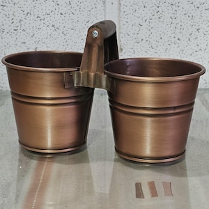 Peut inclure: Un double seau en métal de couleur cuivre avec une poignée en bois. Les seaux sont cylindriques avec un design strié et sont reliés par un support métallique. La poignée est fixée avec une vis.