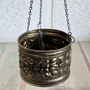 May include: A decorative metal hanging container with a floral pattern. The container is suspended by three black chains. The container has a cylindrical shape with embossed roses and leaves around the exterior.