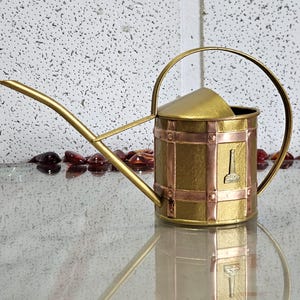 Puede incluir: Una regadera de latón de estilo vintage con un pico largo y un asa arqueada. La regadera presenta detalles en cobre y un pequeño emblema decorativo. El diseño recuerda a las herramientas de jardín antiguas, apta para uso interior o exterior.