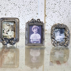 Puede incluir: Tres marcos de fotos plateados antiguos, cada uno con un retrato en blanco y negro de una niña. Los marcos tienen diseños ornamentados, incluyendo cuerdas, lazos y detalles florales. Los marcos se exhiben sobre una superficie reflectante.