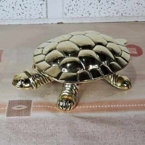 Peut inclure: Une figurine de tortue en métal de couleur dorée avec une carapace détaillée et quatre pattes. La tortue est posée sur une surface brun clair.
