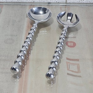 Könnte beinhalten: Ein Set aus zwei silbernen Salatbesteck mit Perlengriffen. Der Servierlöffel und die Gabel sind aus Metall und haben eine polierte Oberfläche.