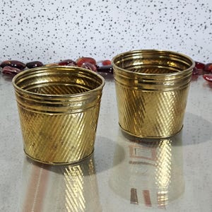 May include: Two gold-coloured metal containers with a textured, diagonal pattern. The containers are cylindrical with a slightly flared top and sit on a reflective surface. The background features decorative stones.