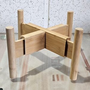 Peut inclure: Un support de plante en bambou naturel avec une base en forme de croix et quatre pieds cylindriques. Le support est conçu pour contenir une plante en pot et a une esthétique simple et moderne. Le support mesure environ 20 cm de haut.