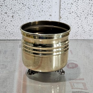 May include: A small, antique brass planter with a round body and three decorative feet. The planter has horizontal bands around the middle and a dark interior. The brass has a polished finish, reflecting light.