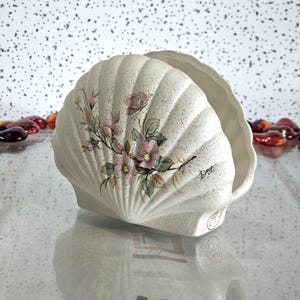 May include: A cream-coloured ceramic seashell-shaped container with a textured surface. It features pink flowers, green leaves, and a butterfly design. The container has a scalloped edge and a small base.