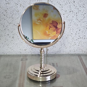 Peut inclure: Un miroir de courtoisie rond de couleur dorée avec un support assorti. Le miroir reflète une peinture avec des images florales orange et jaune. Le support a une base décorative à plusieurs niveaux et un support vertical mince.