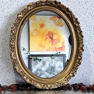 Peut inclure: Un miroir ovale doré avec un cadre orné et texturé. Le miroir reflète une peinture avec des images florales jaunes et oranges et une impression florale en noir et blanc. Des perles de verre brun-rouge sont éparpillées à la base.