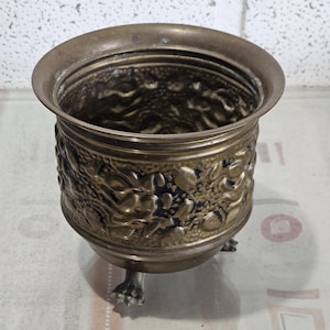 Könnte beinhalten: Ein Messing-Pflanztopf mit einem verzierten Blumenmuster und drei Füßen. Der Topf ist leer und hat einen abgenutzten, antiken Look.
