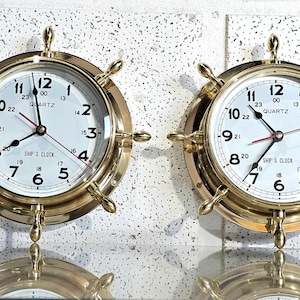 Op de afbeelding: Twee messing scheepsklokken met witte wijzerplaten en zwarte cijfers. De klokken hebben een quartz uurwerk en zijn omgeven door een messing frame met een scheepsradontwerp. De tekst "SHIP'S CLOCK" is zichtbaar.