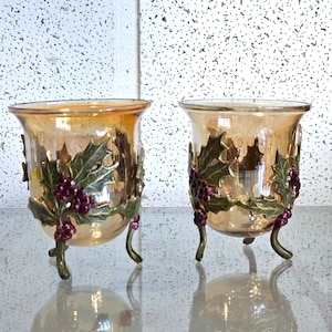 Pode incluir: Dois castiçais de vidro âmbar com detalhes de folhas de azevinho e bagas em metal dourado. Os castiçais têm três pernas e estão sobre uma superfície branca.