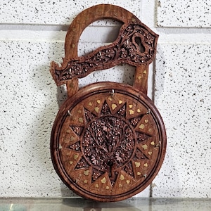 Peut inclure: Un cadenas décoratif en bois avec des sculptures complexes. La base circulaire présente un motif floral et en étoile détaillé, orné d'incrustations en laiton. La poignée et la partie supérieure sont également sculptées de motifs floraux.