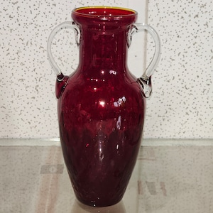 Op de afbeelding: Een donkerrode glazen vaas met twee heldere glazen handvatten. De vaas heeft een getextureerd oppervlak en een licht uitlopende opening.