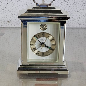 May include: Silver-toned alarm clock with a white face and black hands. The clock has Roman numeral hour markers and a gold-coloured second hand. The clock face also has the words "Kenox" and "Quartz Alarm".