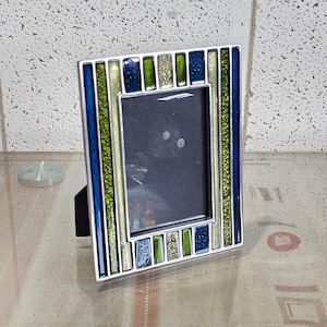 Könnte beinhalten: Ein dekorativer silberner Bilderrahmen mit rechteckiger Öffnung. Der Rahmen ist mit blauen, grünen und silberfarbenen Glaseinsätzen verziert. Er hat eine schwarze Staffelei-Rückseite für die Tischpräsentation.