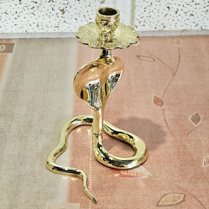 May include: A gold metal candle holder shaped like a cobra with a decorative floral design on the top. The candle holder is on a pink and white patterned surface.