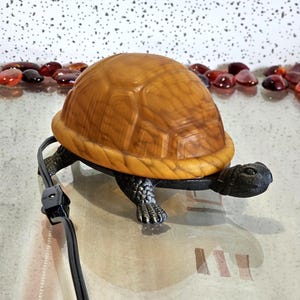 May include: A turtle-shaped lamp with a brown shell and black base and legs. The shell has a textured pattern. The lamp has a black power cord and plug. The turtle's head is facing right.