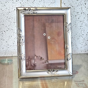 May include: A silver picture frame with a floral design. The frame has a wood backing and a glass front. The frame is sitting on a table.