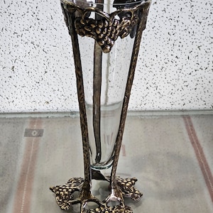 Może przedstawiać: Przezroczysty szklany wazon na pojedyncze kwiaty z ozdobną złotą metalową ramą w kształcie winorośli. Wazon ma okrągłą podstawę i wysoki, smukły kształt.